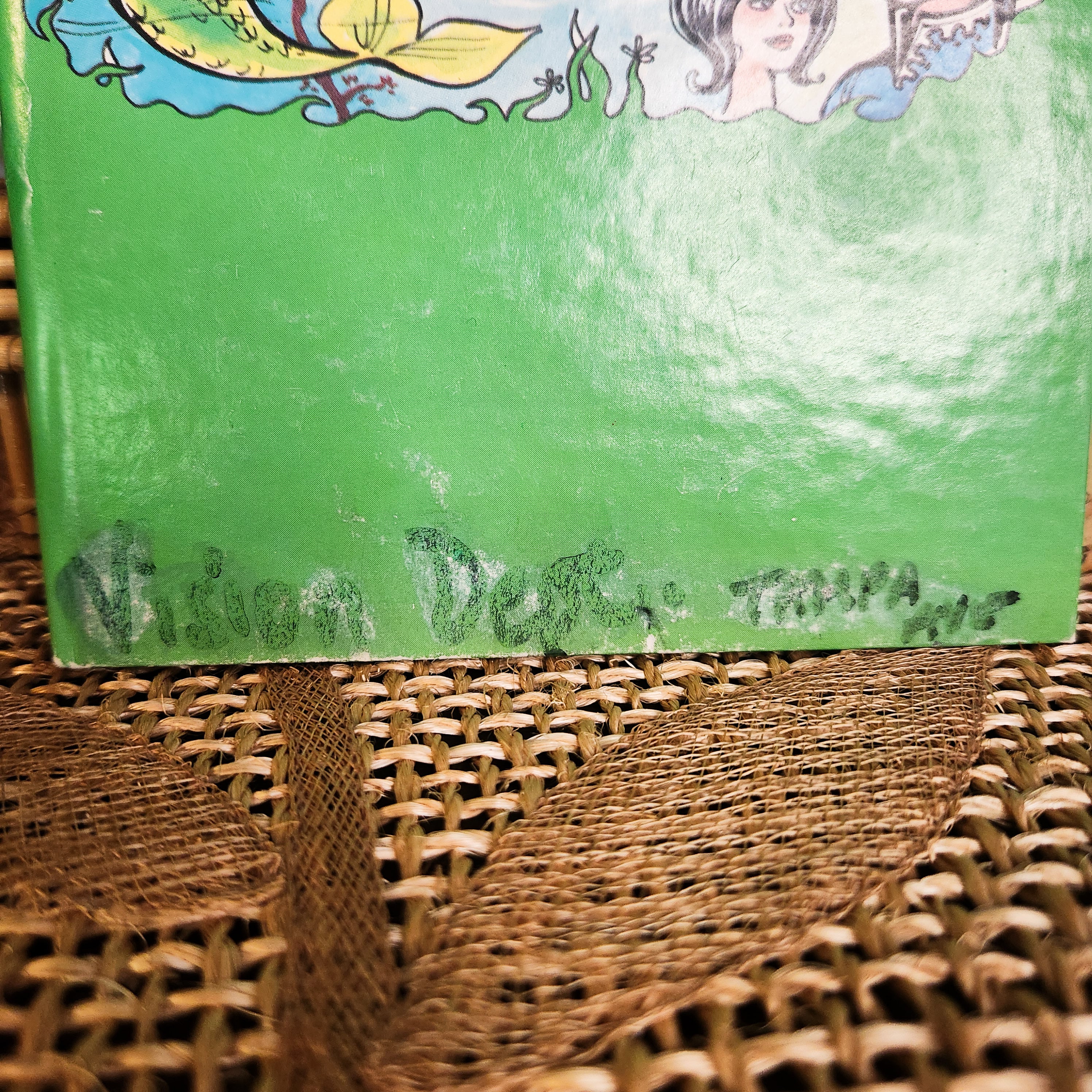 Green book with a colorful top edge on a woven surface with a name written in sharpie that has been wiped down, but was not removed